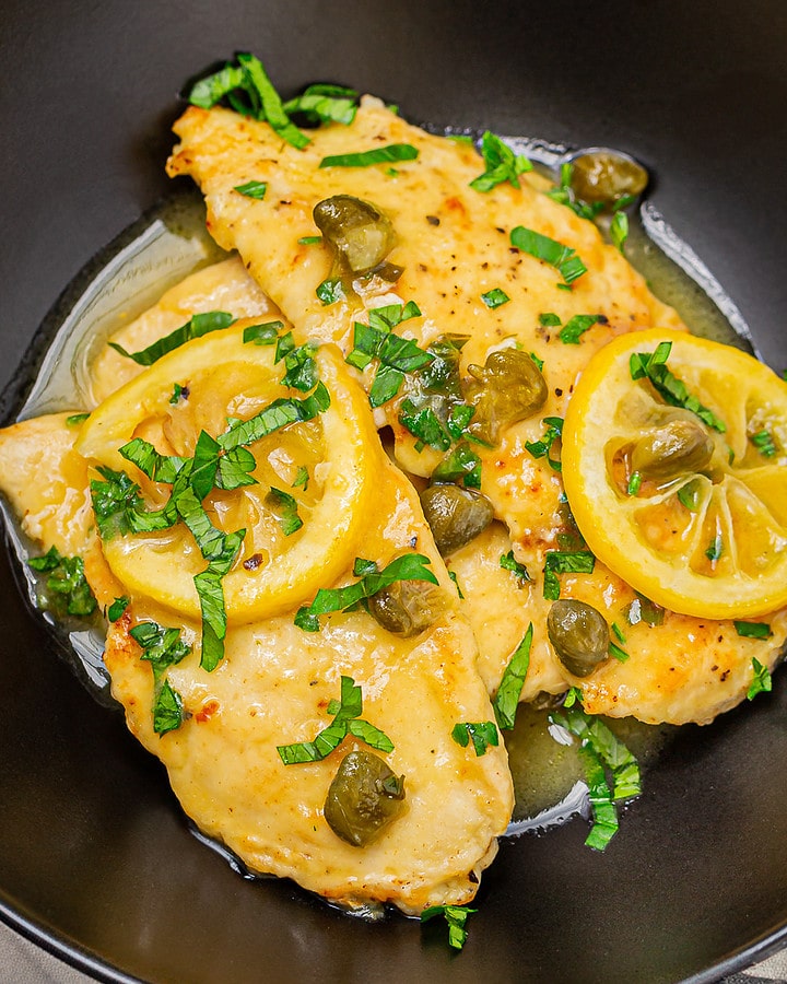A vibrant dish of chicken piccata, featuring golden-browned chicken breasts topped with lemon slices and capers, garnished with chopped parsley, presented in a dark bowl on a grey background.