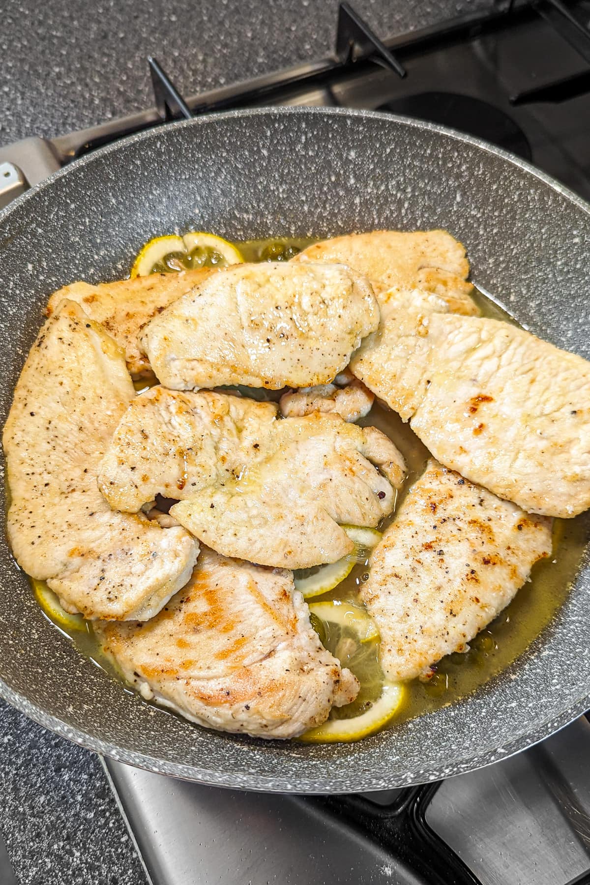 Sautéed chicken breasts in a frying pan, cooked to a golden-brown sear, bathed in a lemony sauce with slices of lemon.