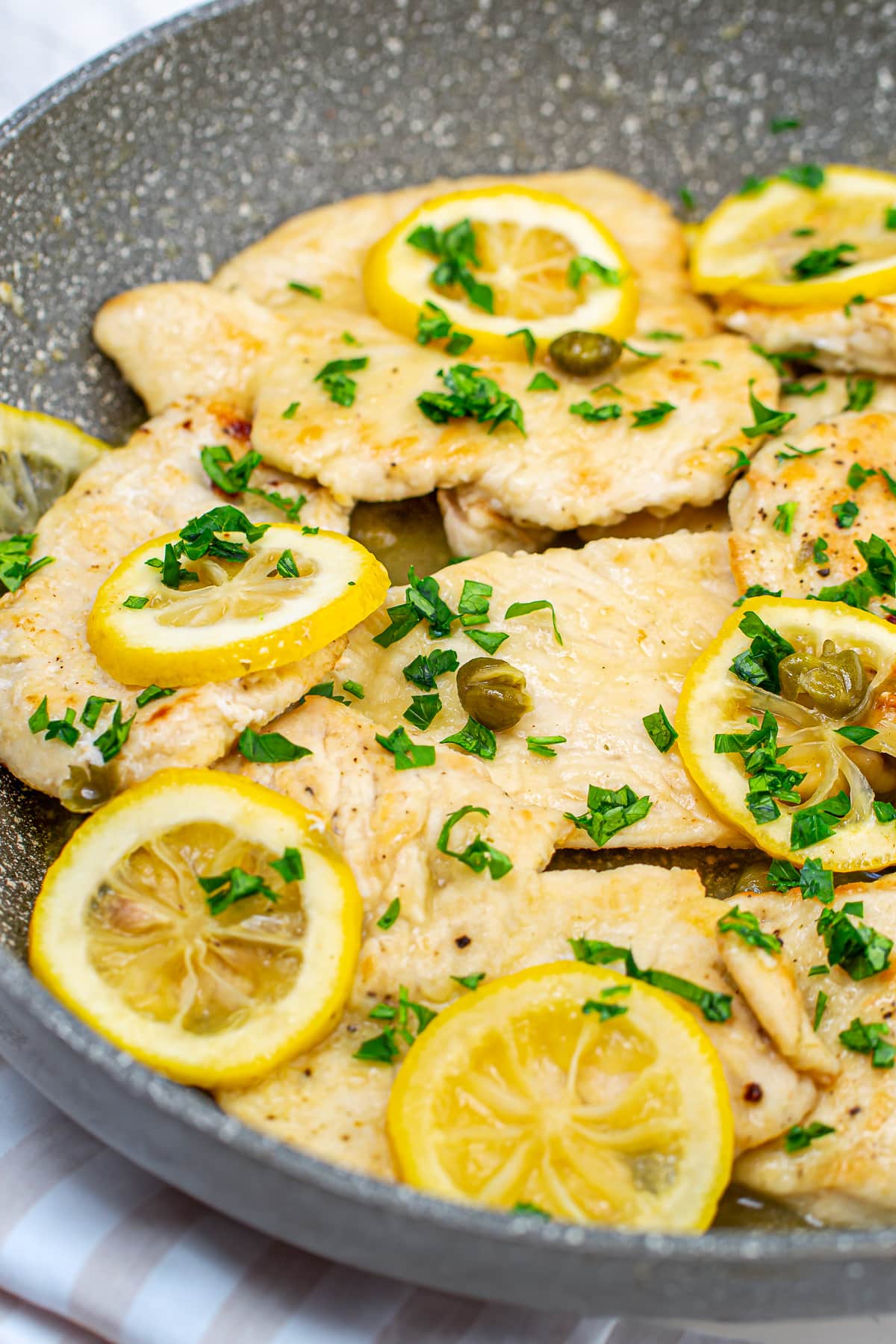 Close-up of chicken piccata in a frying pan, topped with lemon slices and capers, garnished with chopped parsley, ready to be served.