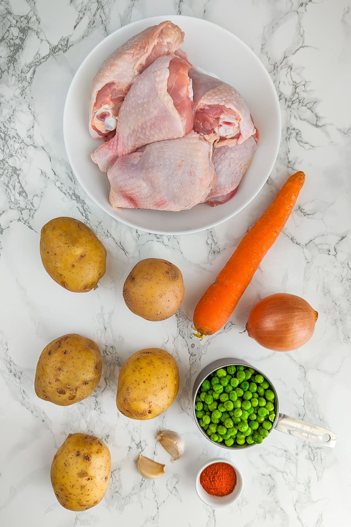 Ingredients for a hearty chicken stew, displayed on a marble surface, including raw chicken legs, potatoes, carrots, onions, peas, garlic cloves, and a bowl of spices.