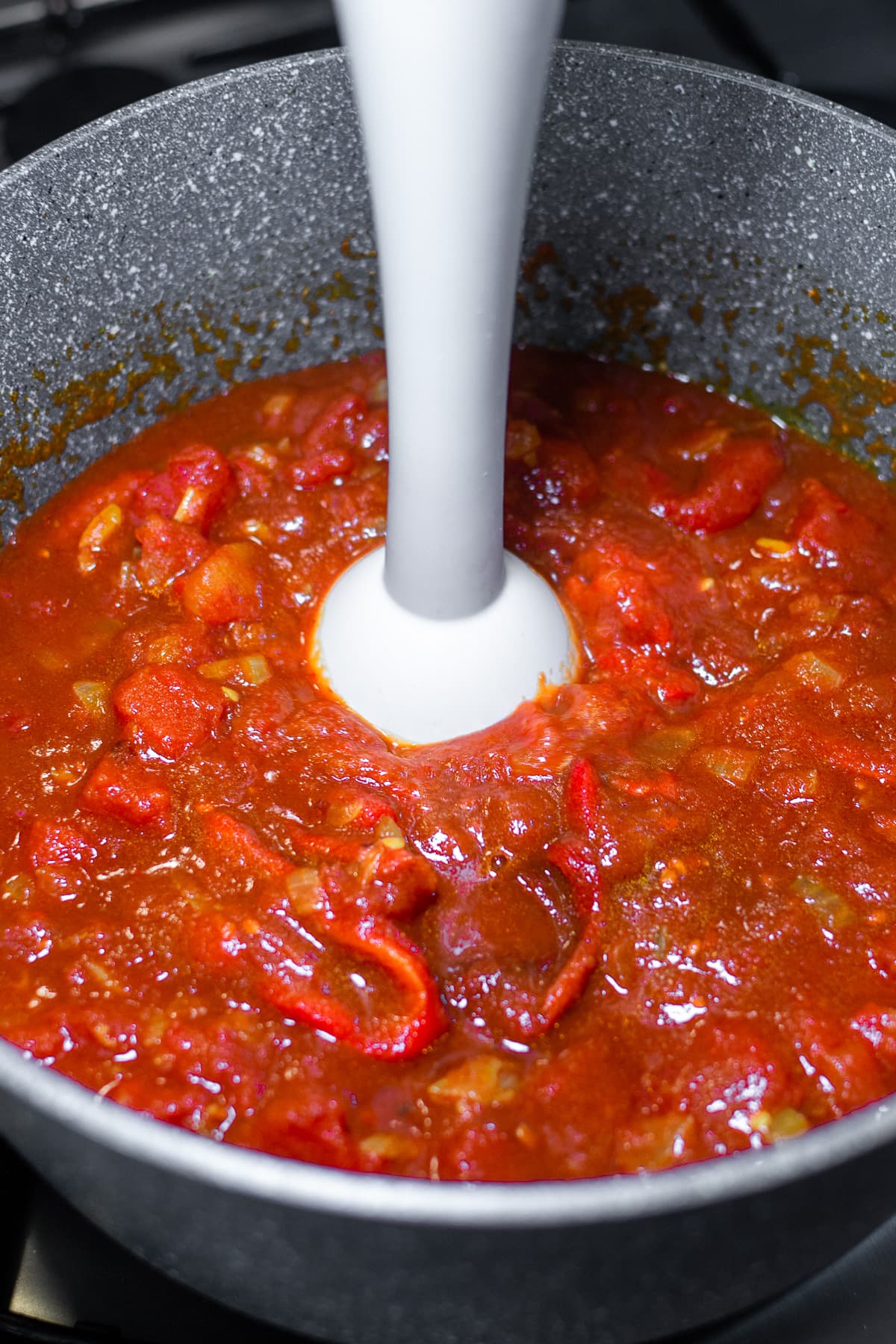 Mixing bell peppers into tomato sauce with a manual white blender.