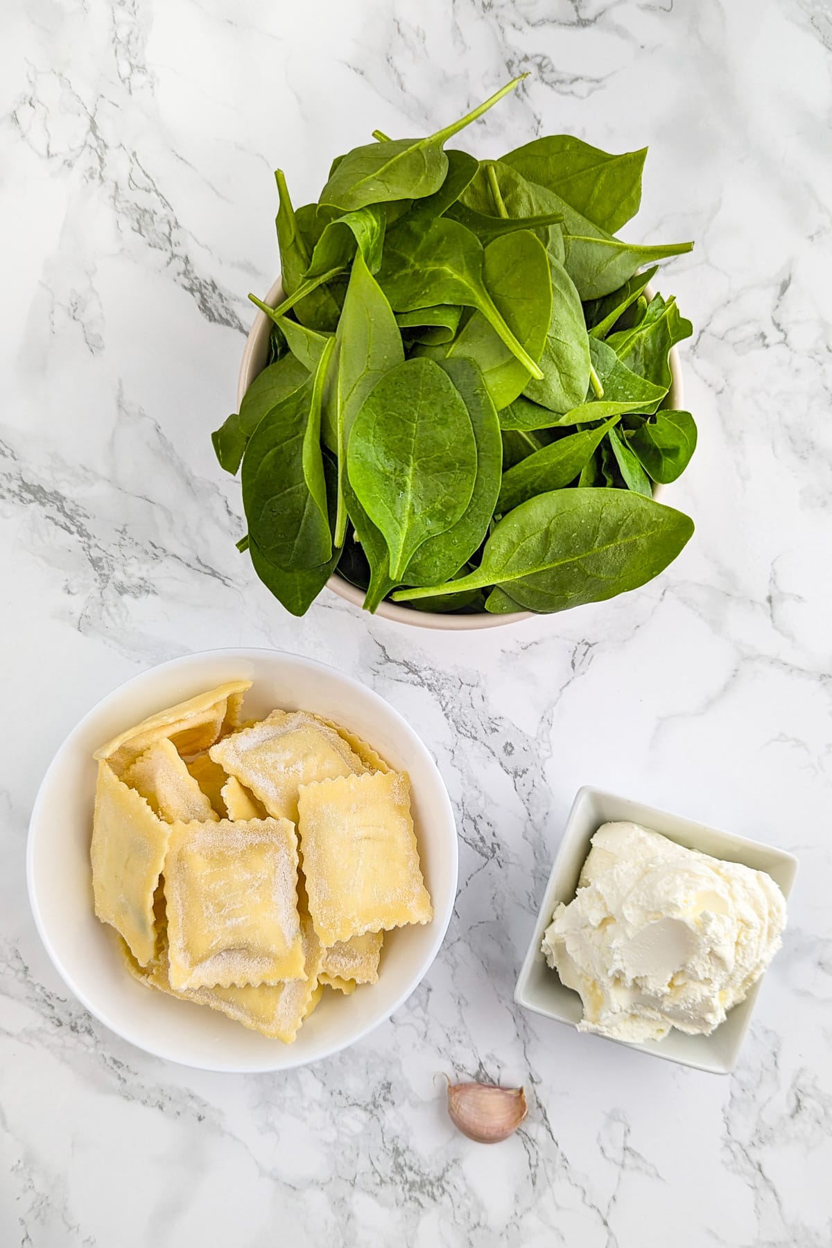 Spinach, cream cheese, garlic and ravioli on a white marble surface.