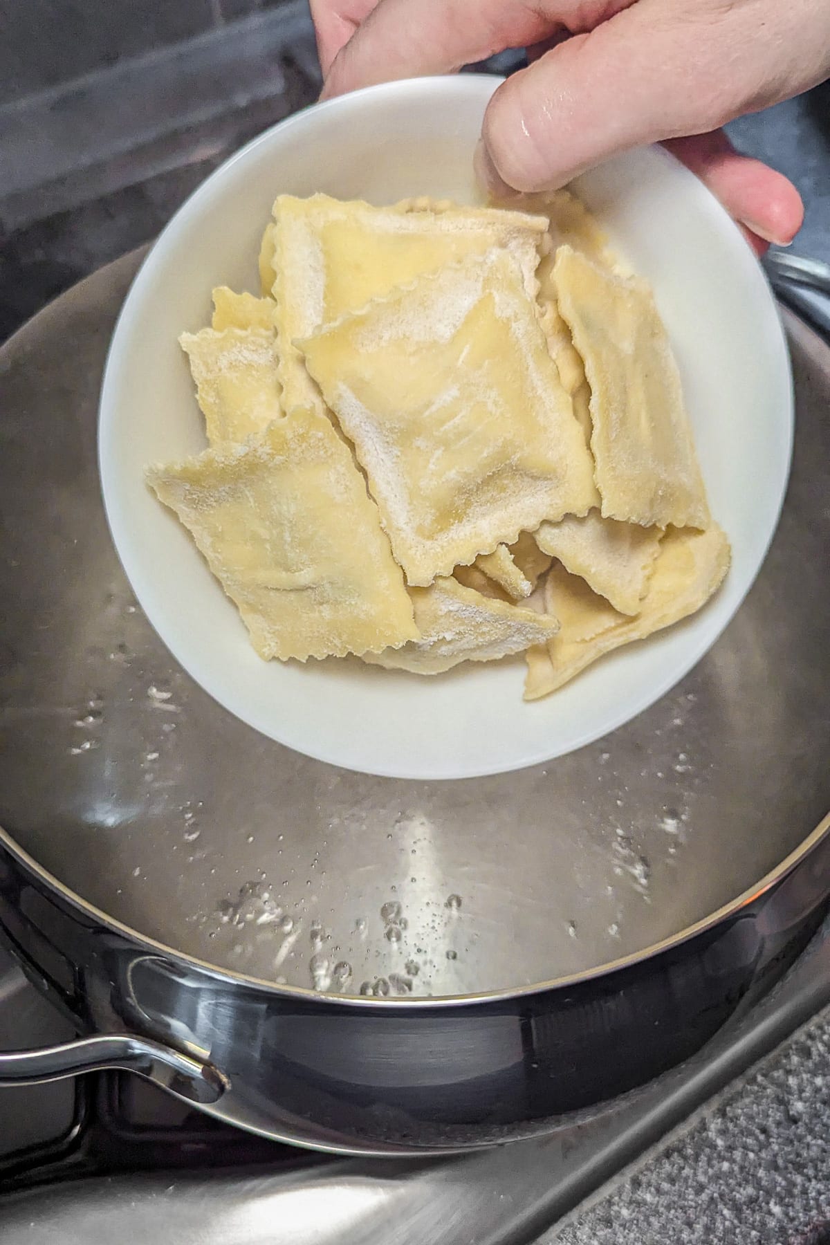 Woman hand holding a plate with raw ravioli over boiling water in a pan.