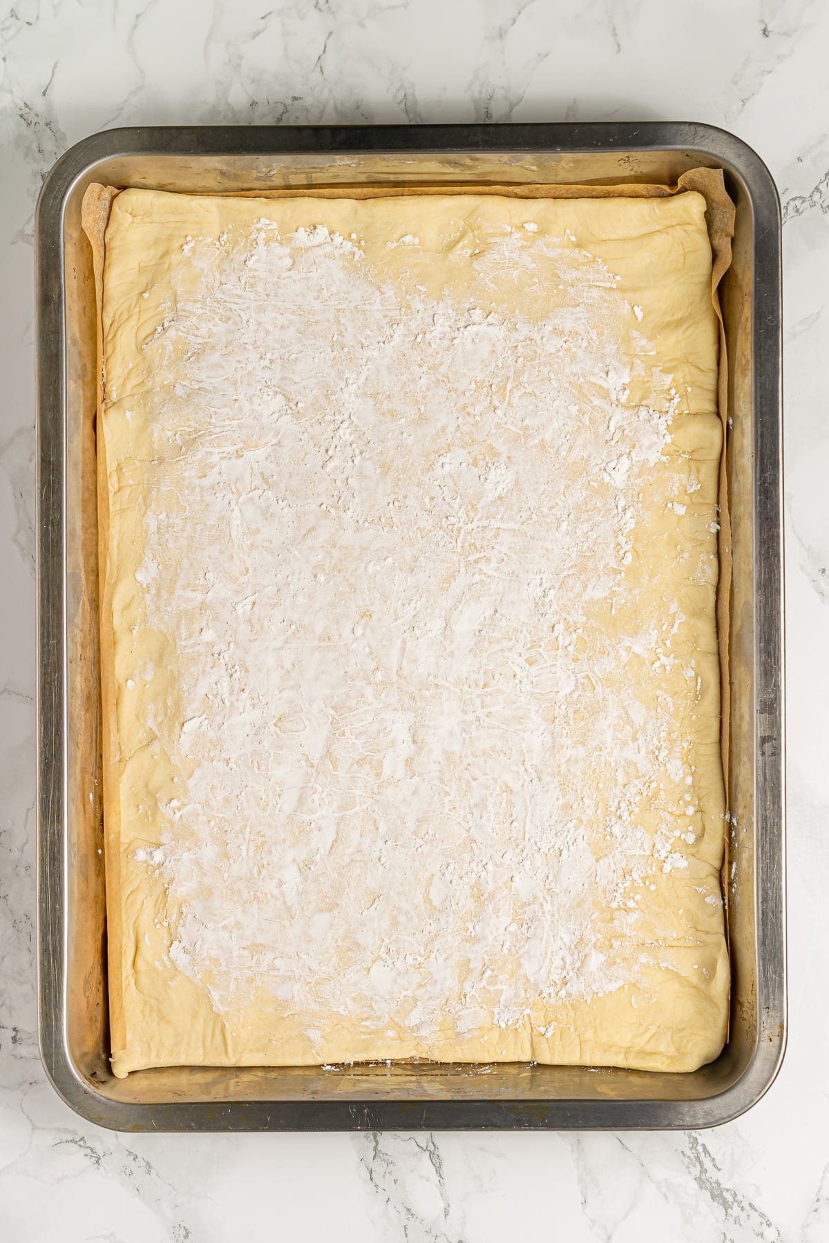 A baking tray lined with parchment paper and unrolled pie dough dusted with flour, ready for assembling an apple tart.