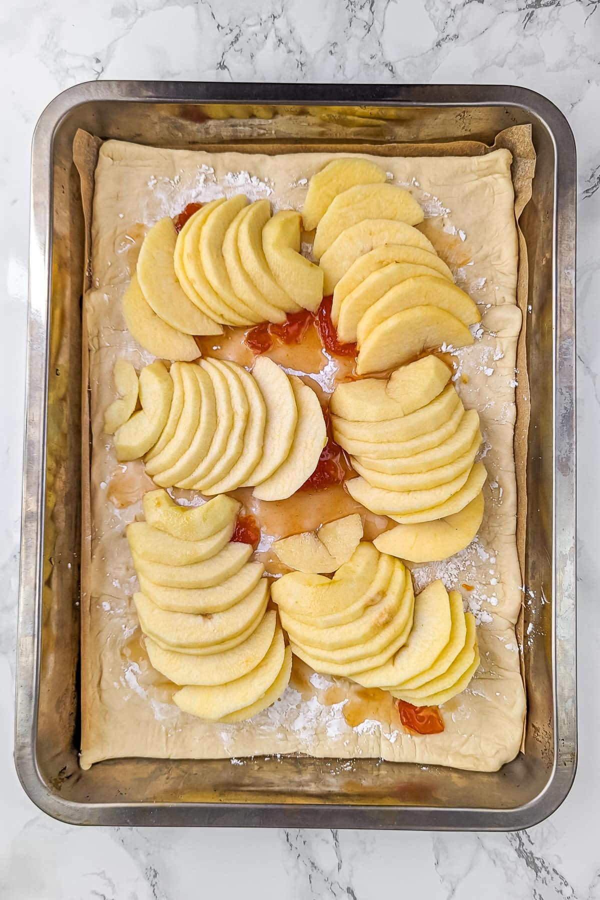 Sliced apples arranged in rows on the pie dough with apricot jam, beginning the tart's assembly.