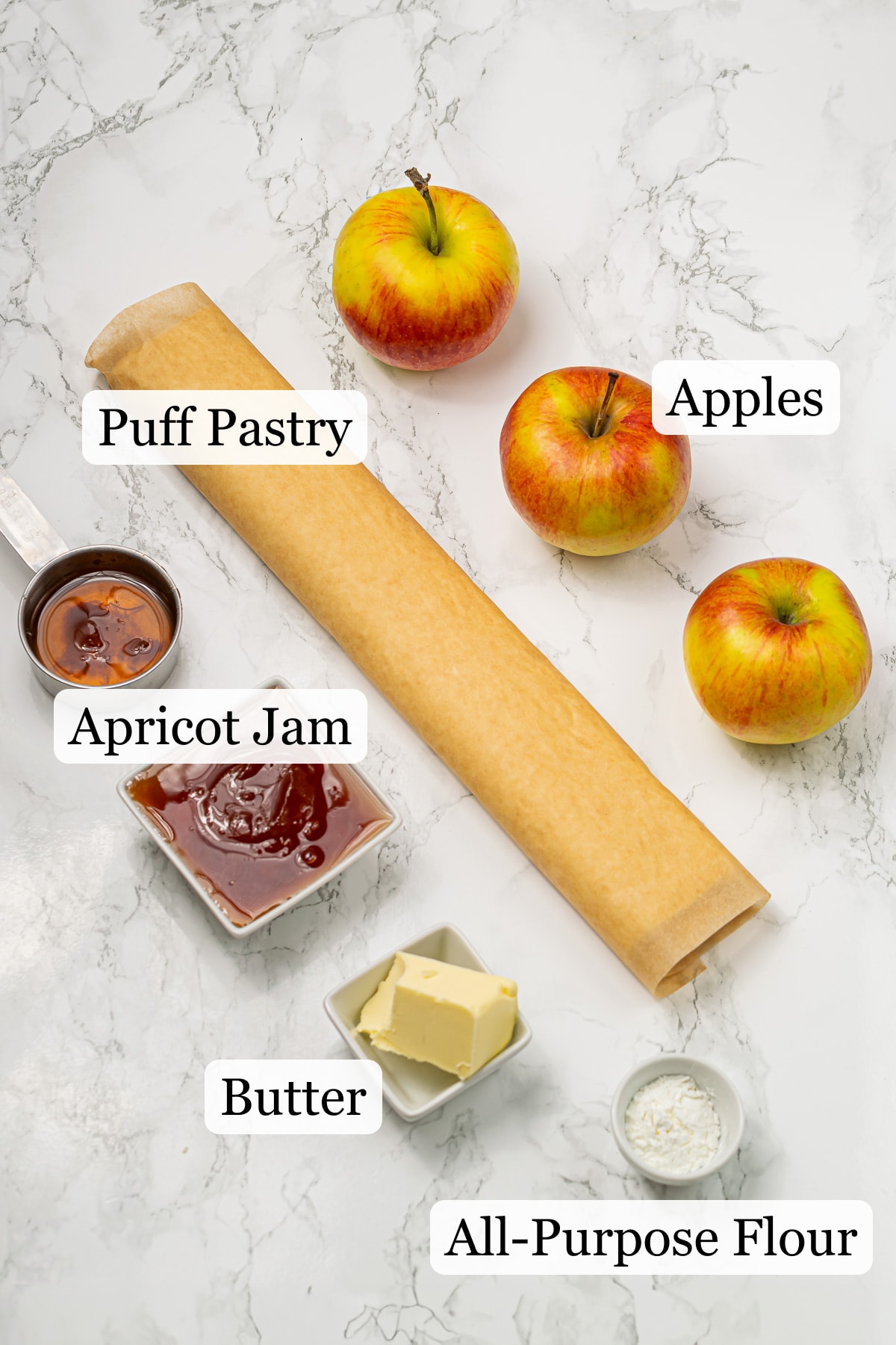 Ingredients for apple tart displayed on a marble countertop, including fresh apples, dough roll, butter, and apricot jam.