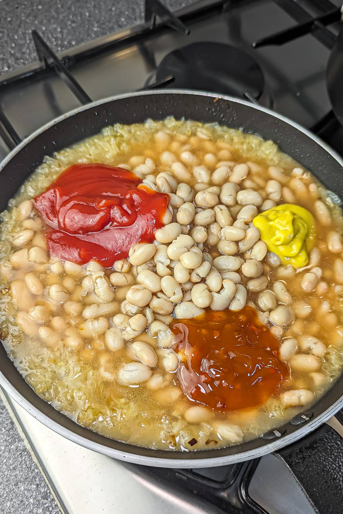 Beans cooking with ketchup, mustard, and chili sauce in a pan.