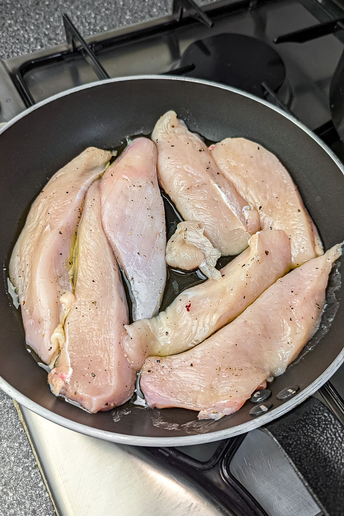 Raw chicken strips seasoned with herbs cooking in a black skillet on a stovetop.