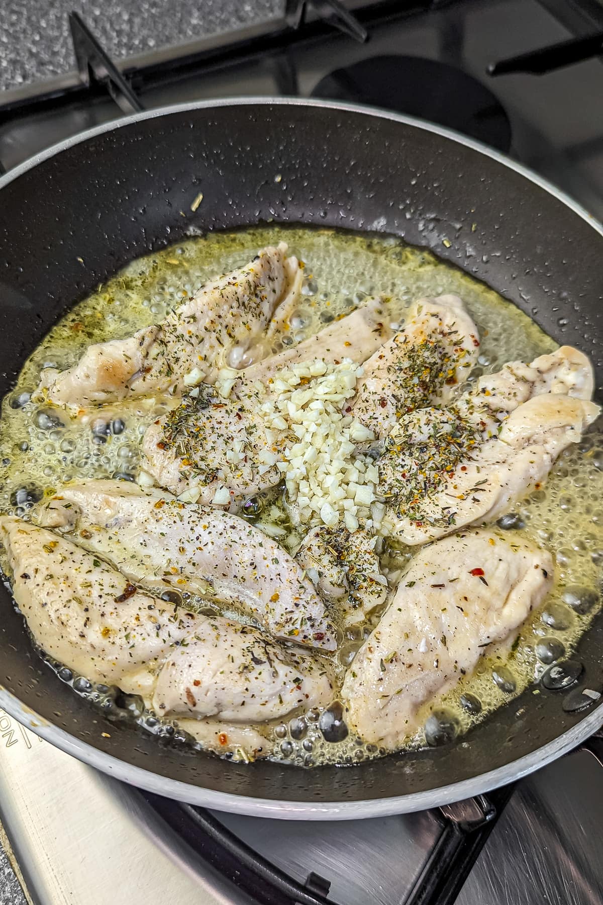 Chicken strips in a skillet being cooked with melted butter, diced garlic, and herbs.