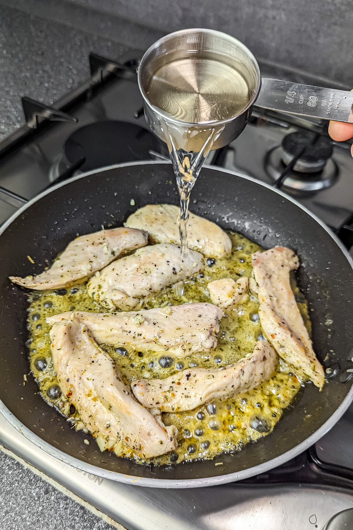 A measuring cup pouring white wine into the skillet with the chicken and herbs.