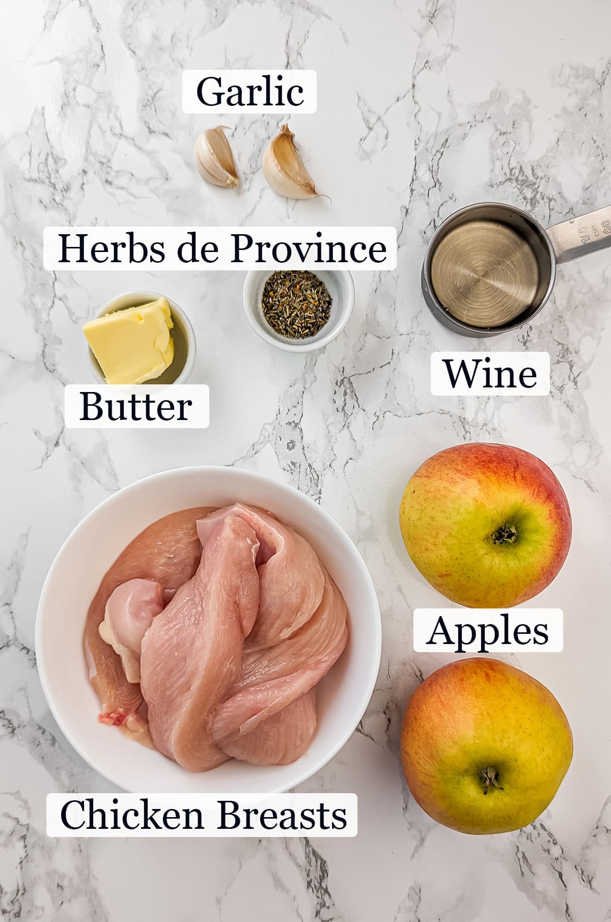 Ingredients for caramelized apple chicken on a marble surface, including raw chicken strips, two whole apples, butter, garlic cloves, herbs, and a measuring cup.