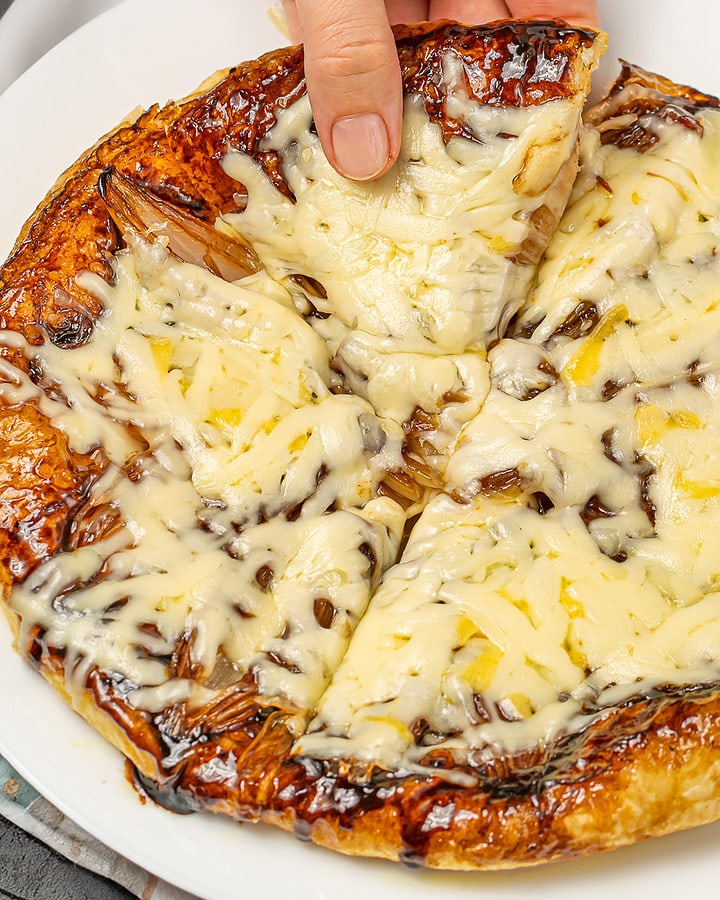 Hand pulling a cheesy slice from a caramelized onion tart, showcasing the melted cheese.