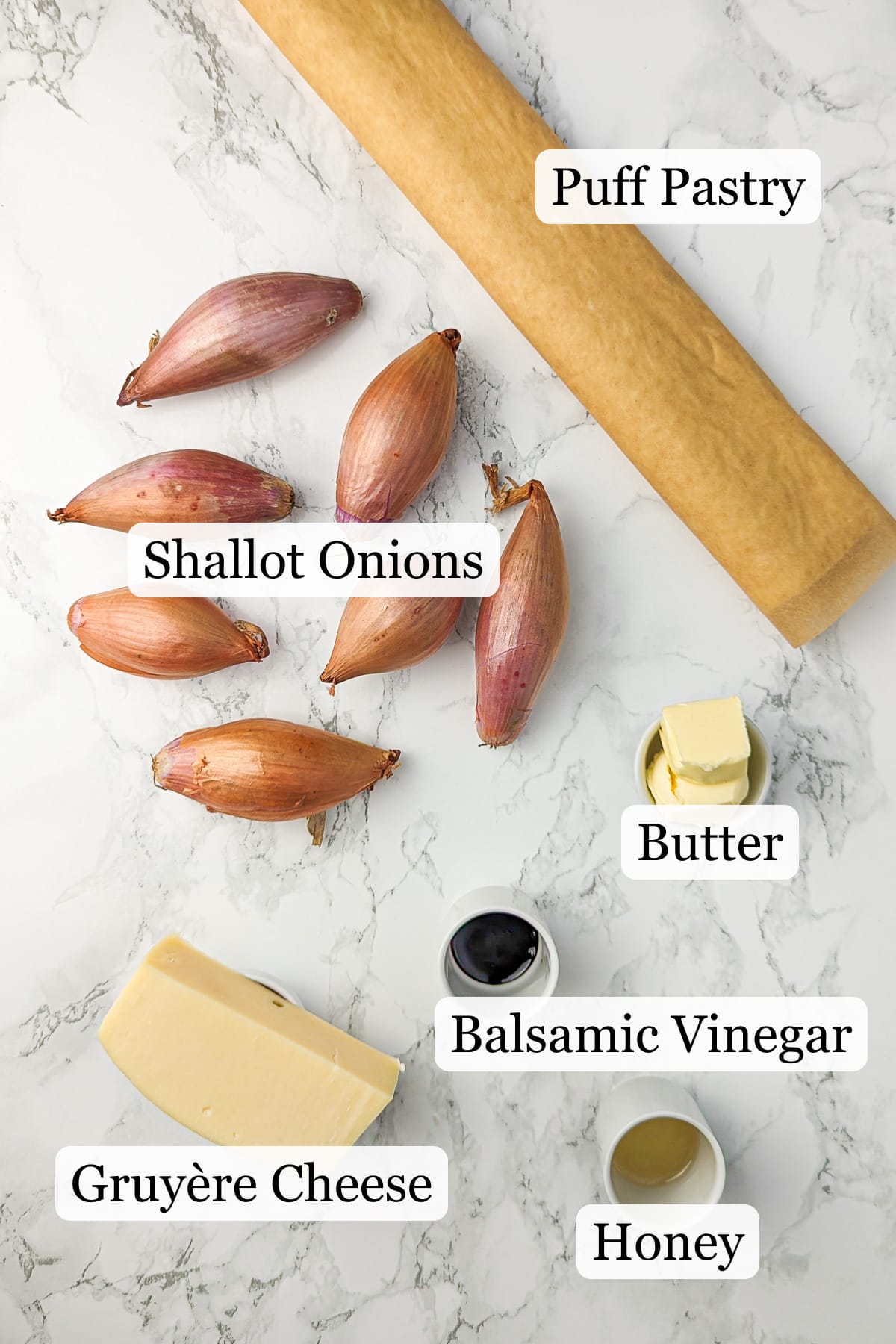 Assorted ingredients for caramelized onion tart on marble background, including shallots, pastry dough, butter, and balsamic vinegar.
