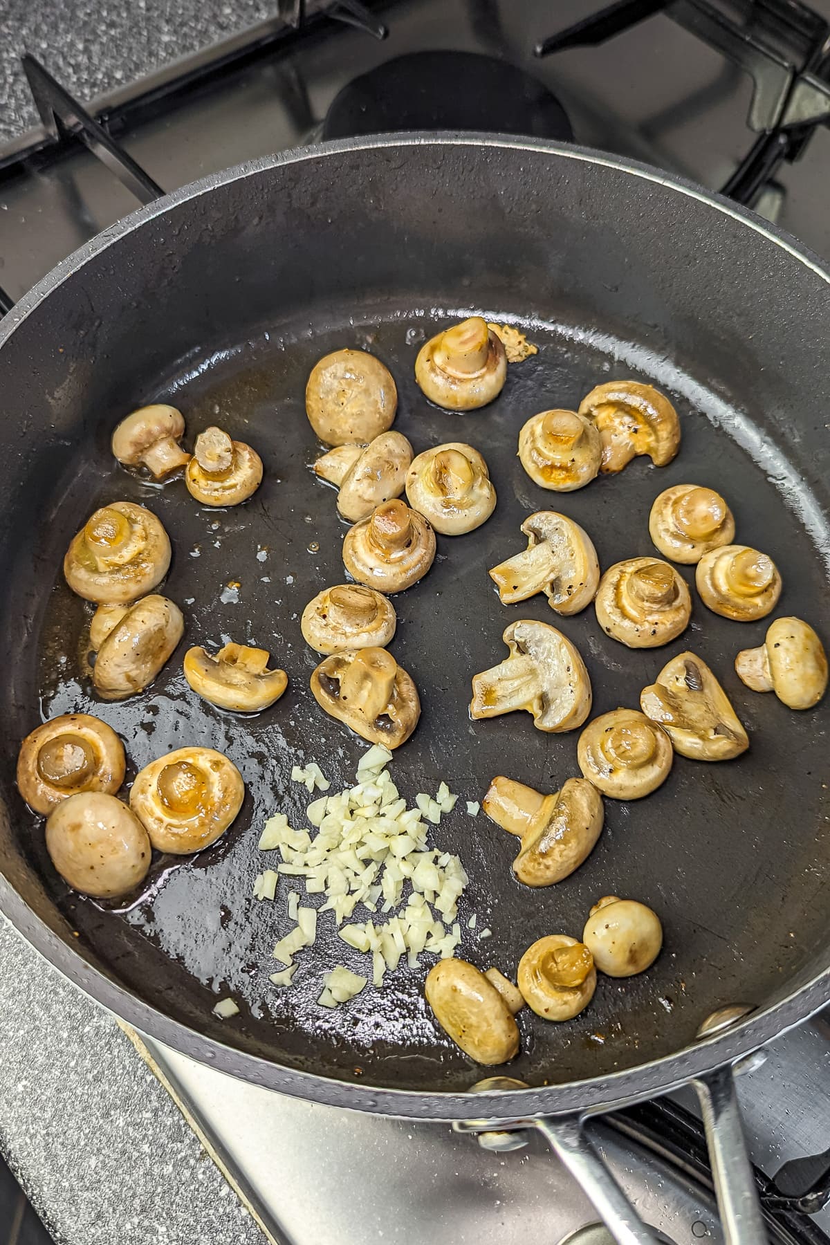 Fresh whole mushrooms being sautéed in a pan with chopped garlic, preparation for a chicken mushroom dish.