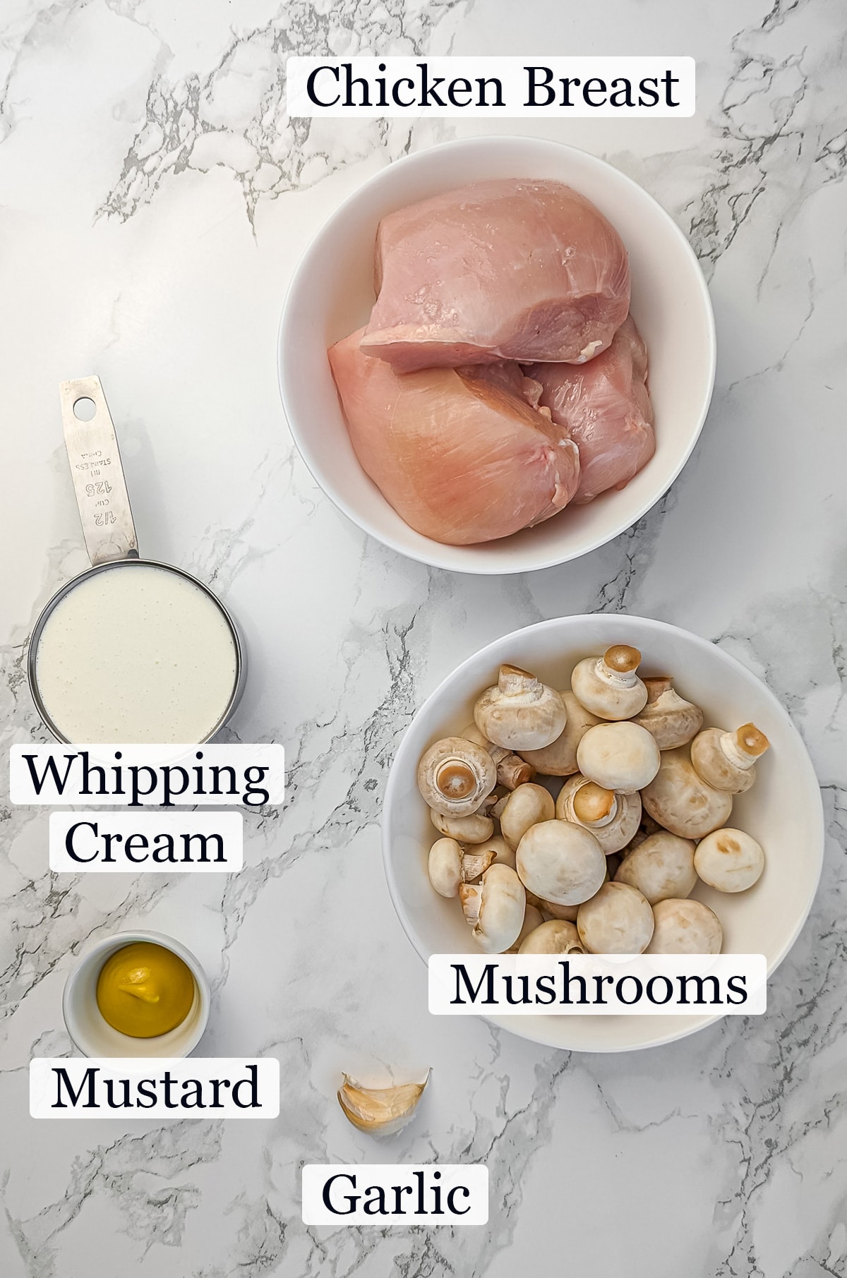 Ingredients for a creamy mushroom sauce recipe on a marble countertop, including chicken breasts, whole mushrooms, heavy cream, olive oil, and garlic.