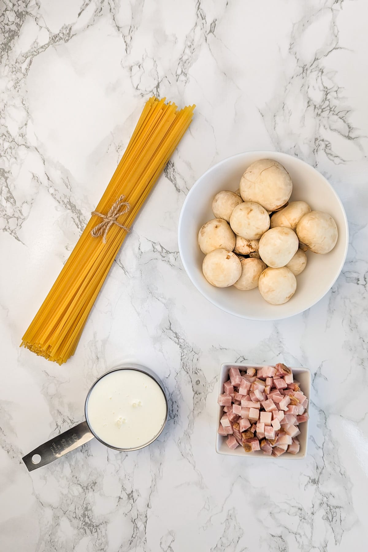 A bundle of uncooked spaghetti tied with twine, a cup of diced ham, a measuring cup of cream, and a bowl of whole mushrooms on a marble countertop.