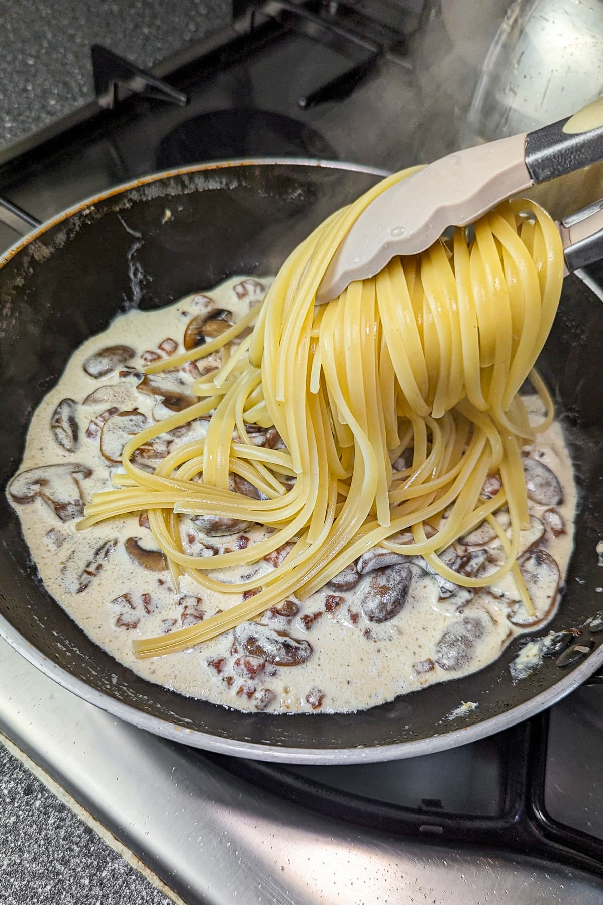 Spaghetti being lifted from the pot with tongs and added to the creamy mushroom and ham sauce in the pan.