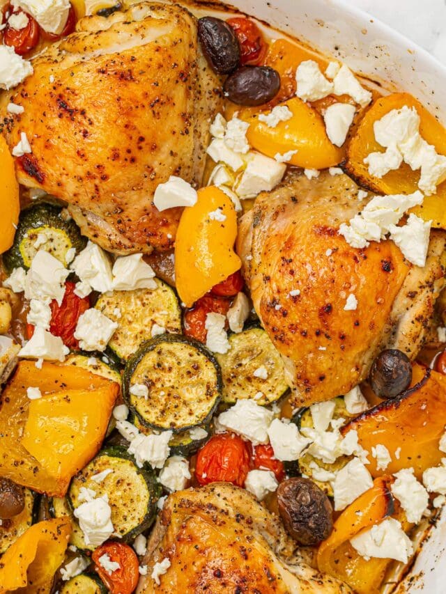 Roasted Greek chicken with bell peppers, tomatoes, zucchini, olives, and crumbled feta cheese in a white baking dish.