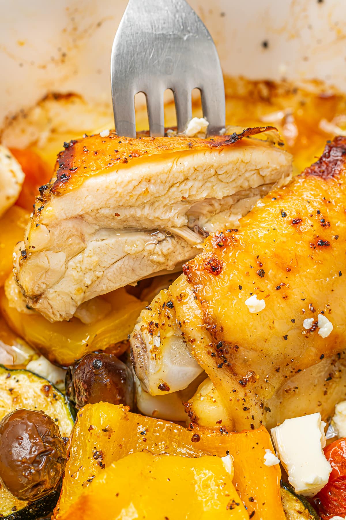 A close-up of a fork cutting into a tender piece of roasted Greek chicken, surrounded by colorful roasted vegetables and feta cheese.
