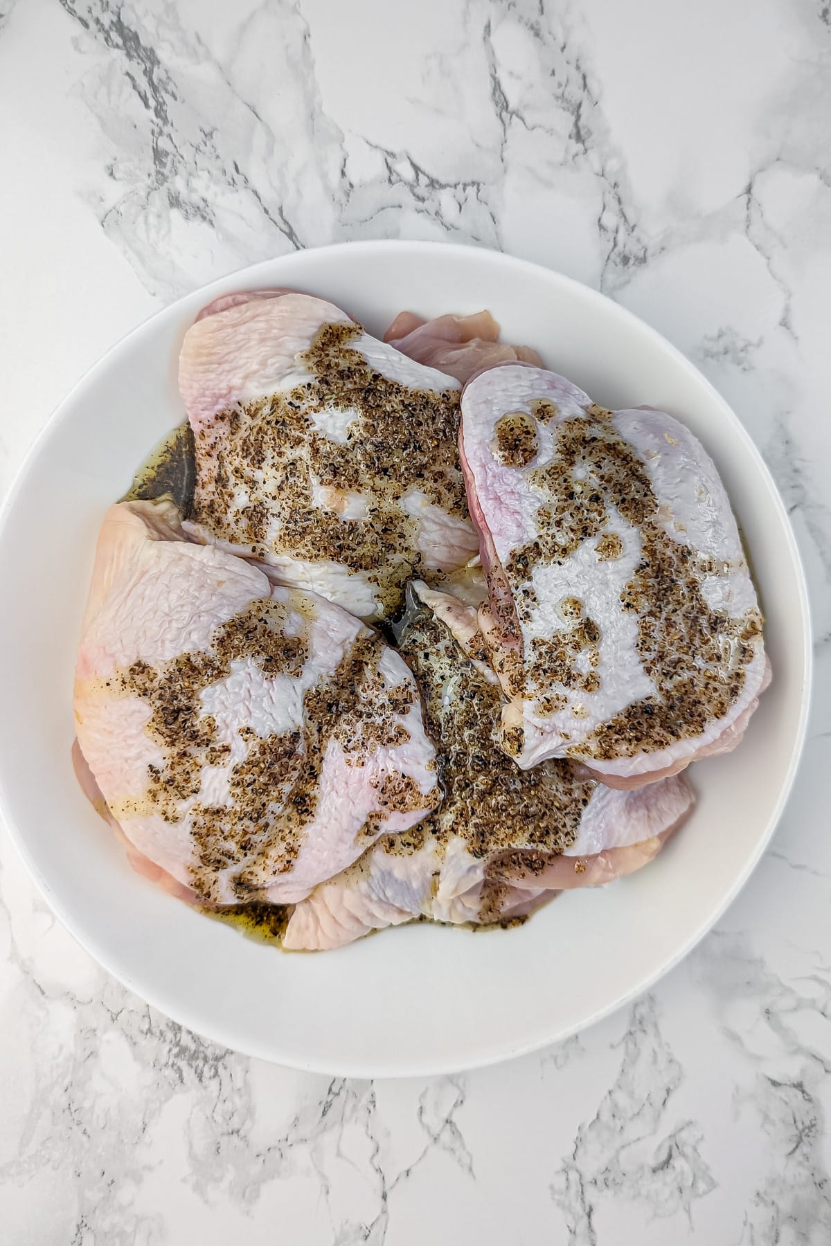 Raw chicken thighs in a white bowl, seasoned with herbs and olive oil for marinating.