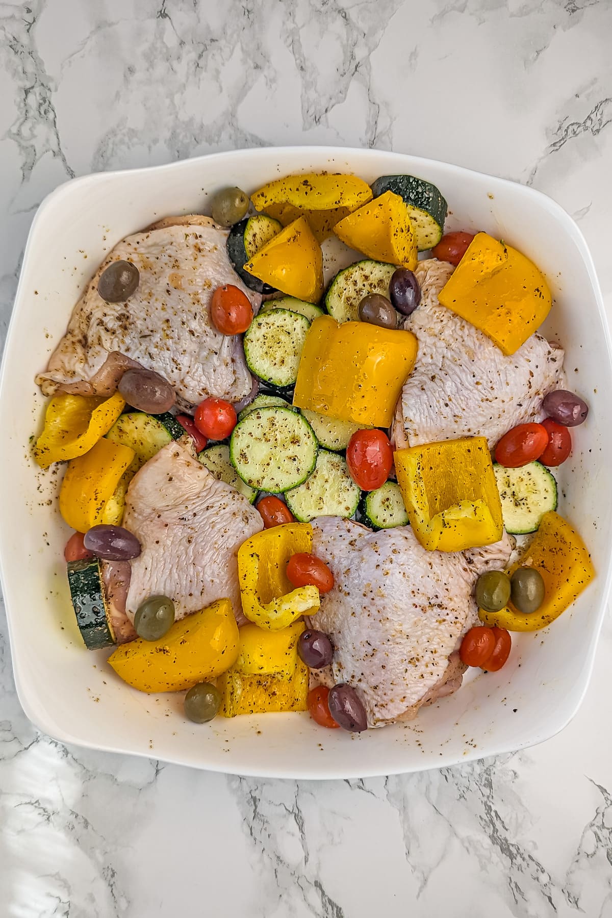Uncooked Greek-style chicken thighs with vegetables and olives arranged in a white baking dish before cooking.