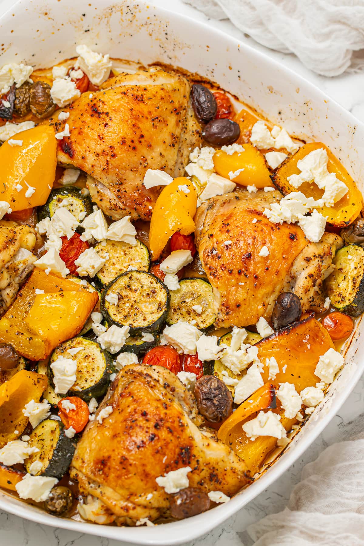 Roasted Greek chicken with bell peppers, tomatoes, zucchini, olives, and crumbled feta cheese in a white baking dish.