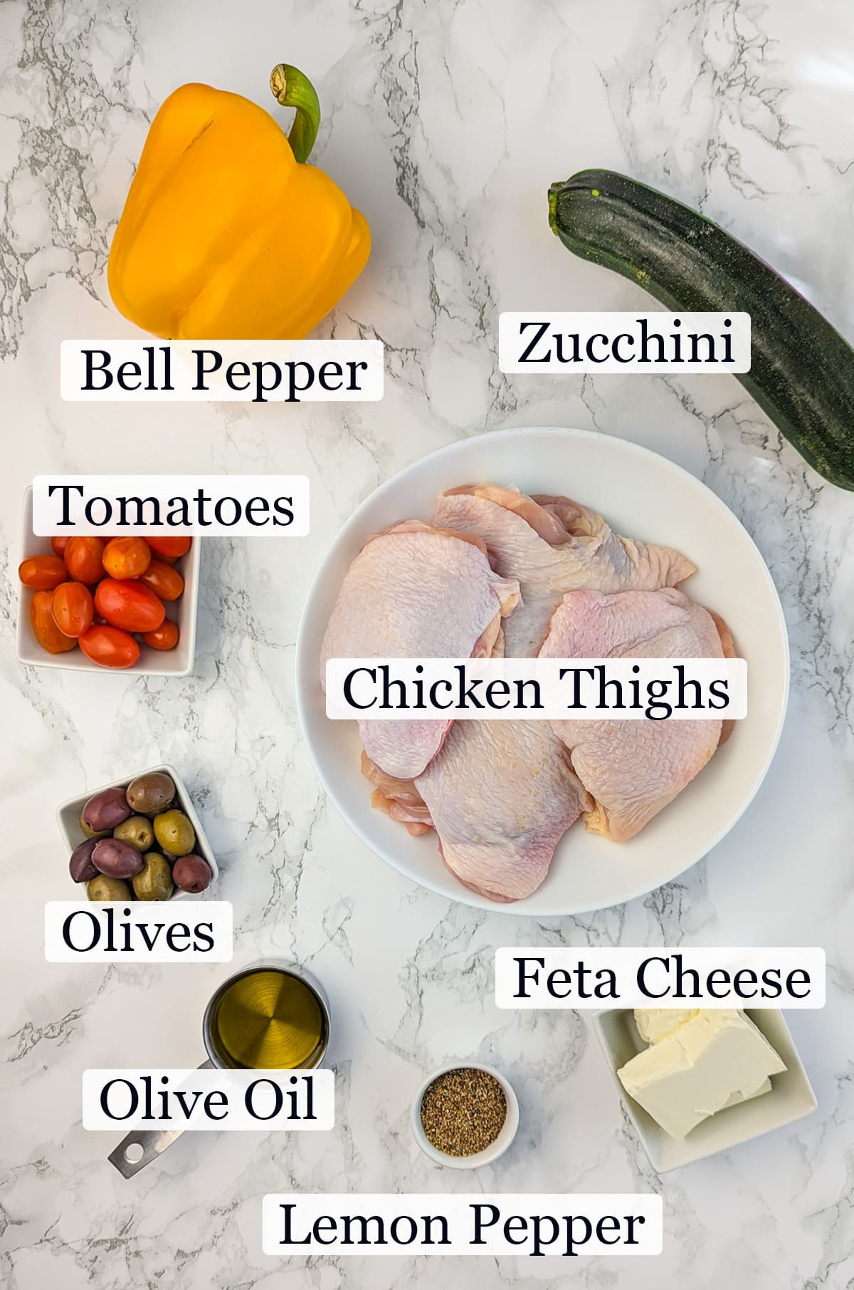 Ingredients for Greek chicken displayed on a marble countertop, including raw chicken thighs, a yellow bell pepper, a zucchini, cherry tomatoes, olives, olive oil, spices, and feta cheese.