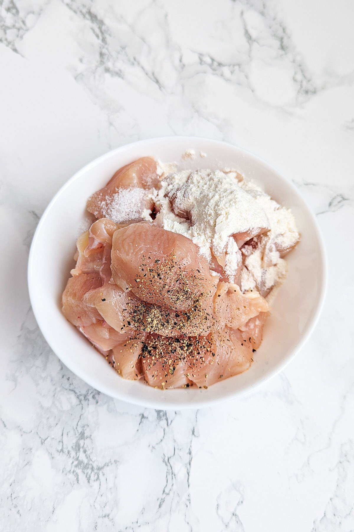 Raw chicken pieces seasoned with salt, pepper, and flour in a white bowl, ready for cooking.