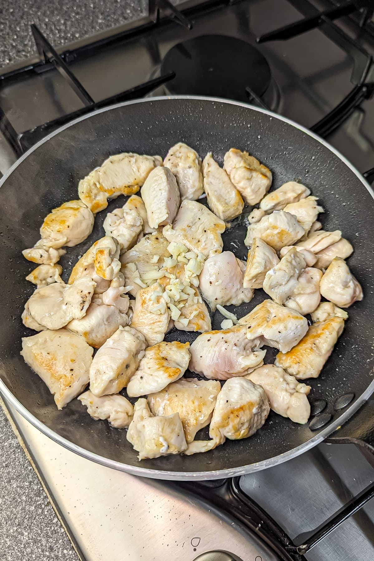 Cooked chicken pieces in a non-stick frying pan with chopped garlic, showcasing the beginning stages of browning.