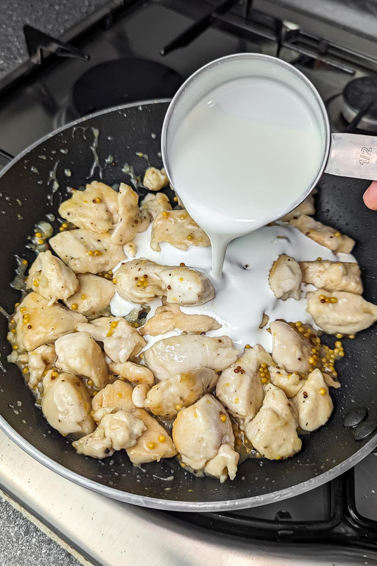 Pouring cream into the frying pan with chicken and mustard, preparing to create a creamy sauce.