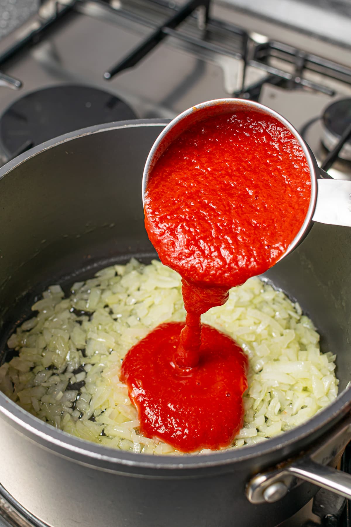 Tomato sauce being poured into a pot of sautéed onions on a stove.