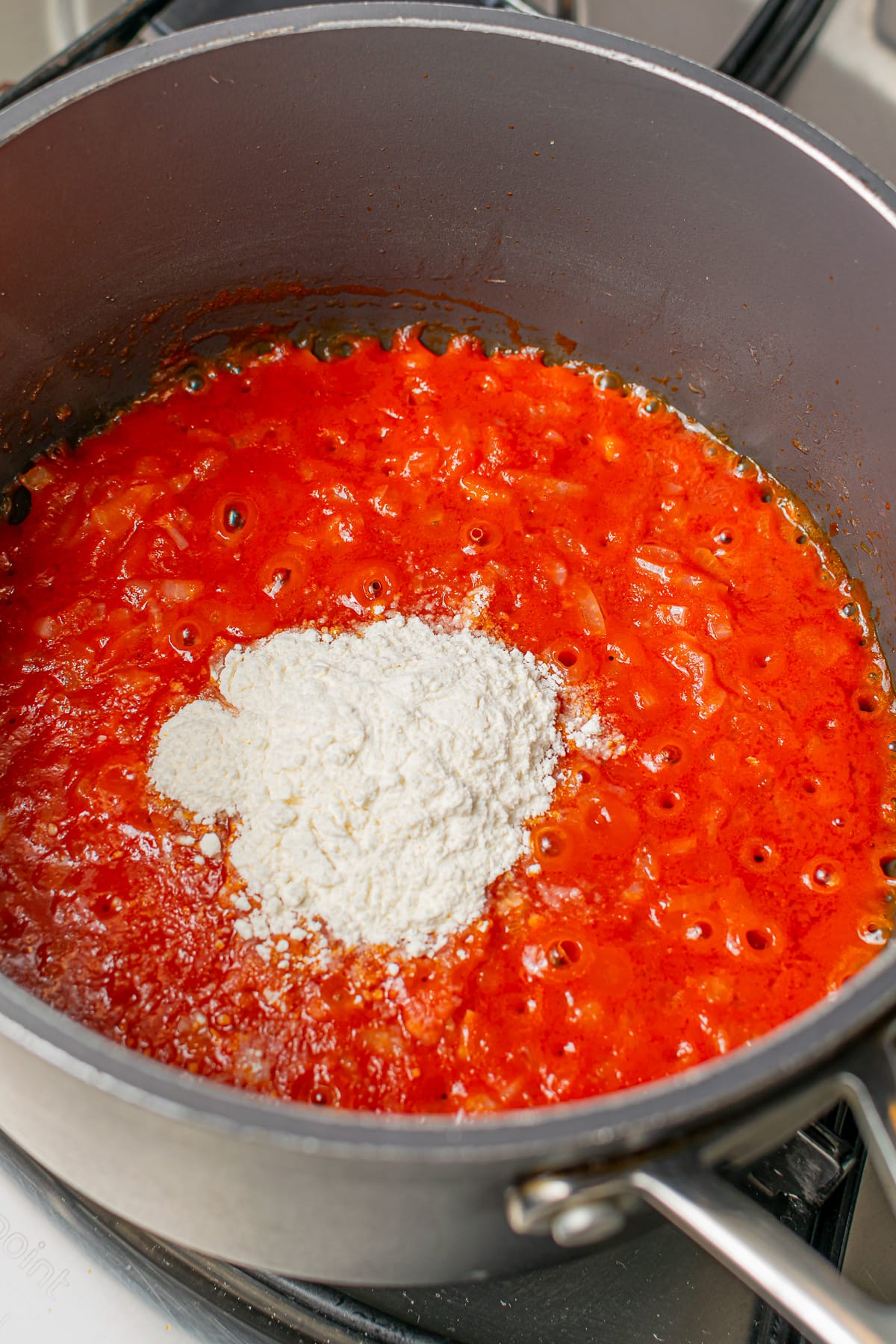 A pot on the stove with tomato sauce and a spoonful of flour added, ready for mixing.