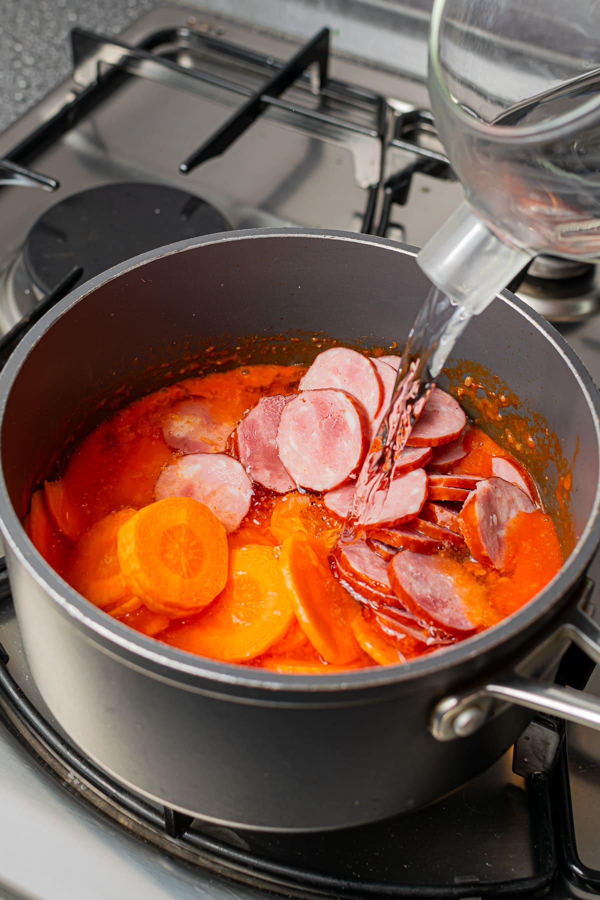 Sliced kielbasa and carrots added to a simmering pot of soup on a stove.