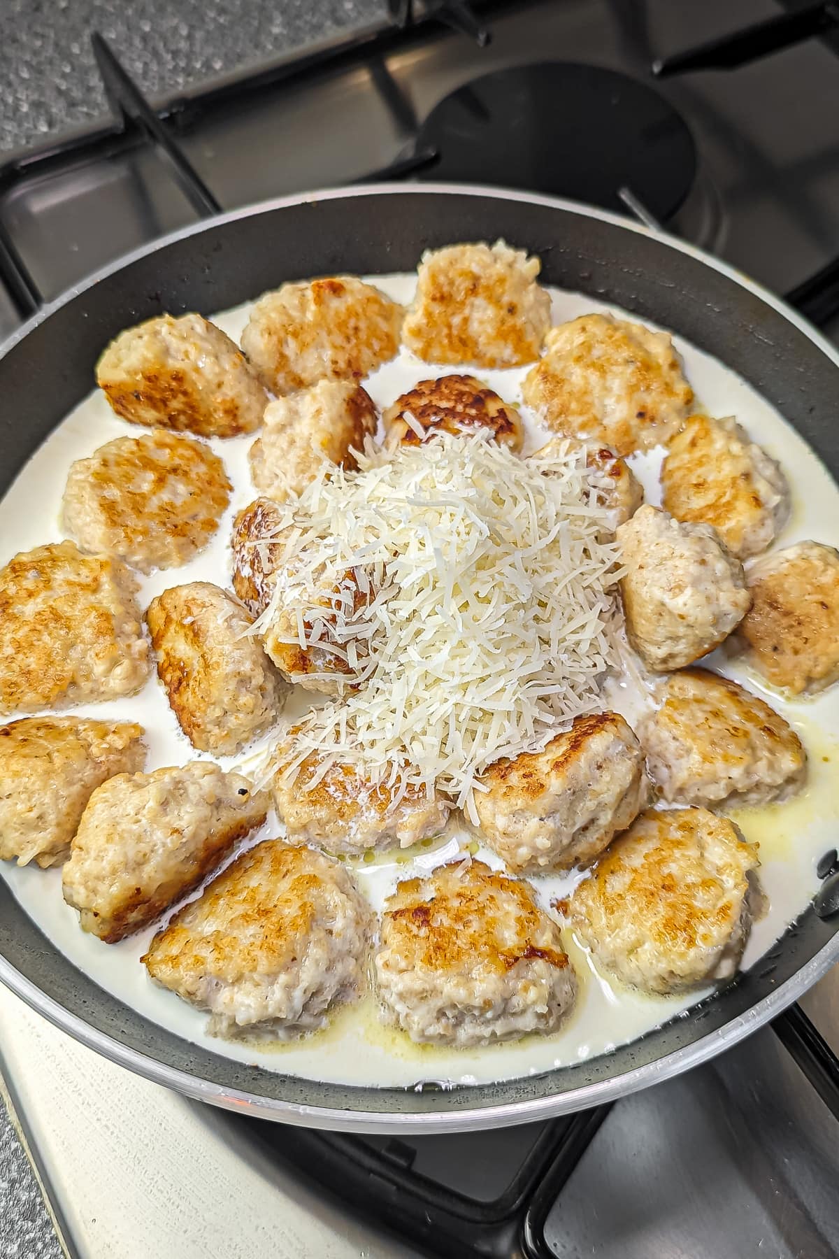 Meatballs in a creamy sauce topped with grated cheese, cooking in a frying pan.