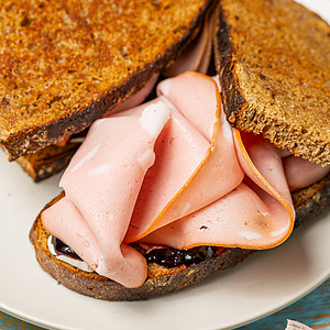 Grilled brown bread sandwich with melted cheese, blackcurrant jam, and folded slices of mortadella on a plate with a blue rim.