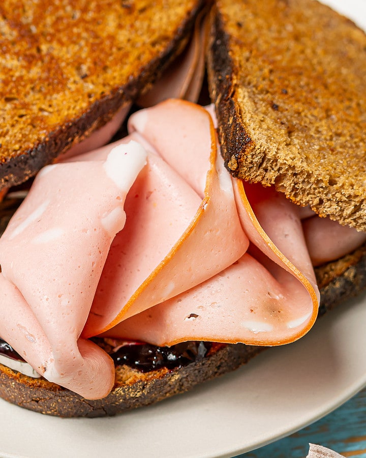 Grilled brown bread sandwich with melted cheese, blackcurrant jam, and folded slices of mortadella on a plate with a blue rim.
