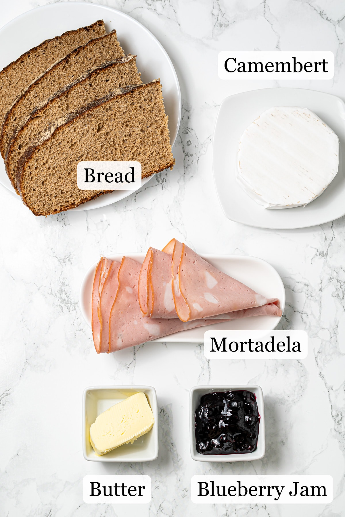 Brown bread slices, soft cheese, mortadella, butter, blackcurrant jam on marble counter.