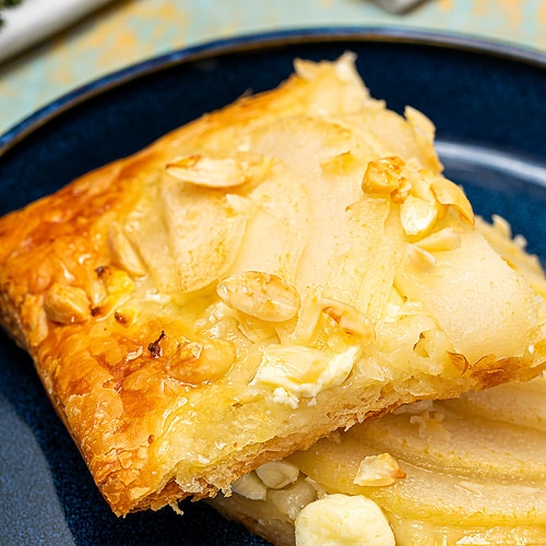 A single slice of pear and feta tart on a blue plate, showcasing the flaky pastry layers and toppings.