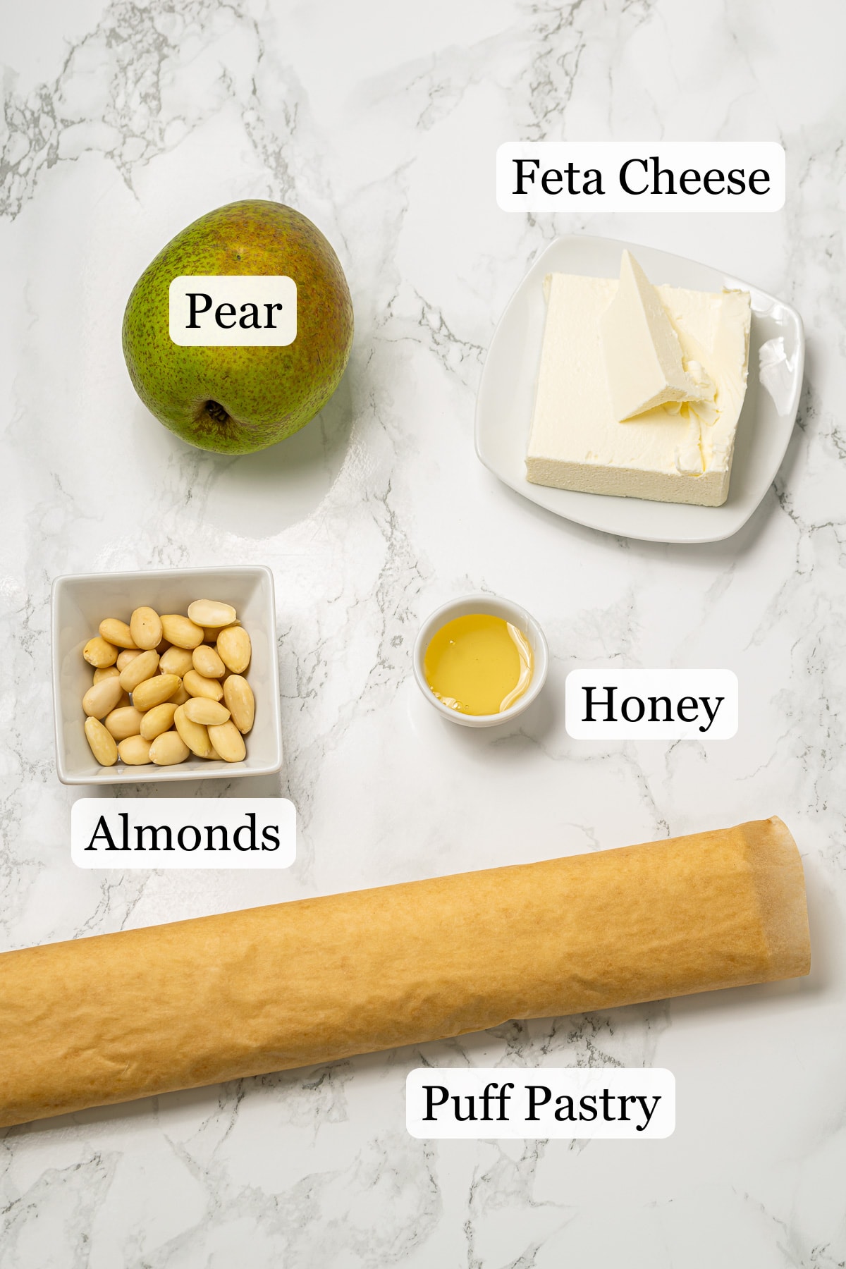 Ingredients for a pear and feta tart on a marble surface, including a whole pear, blocks of feta cheese, almonds, and a rolled dough.