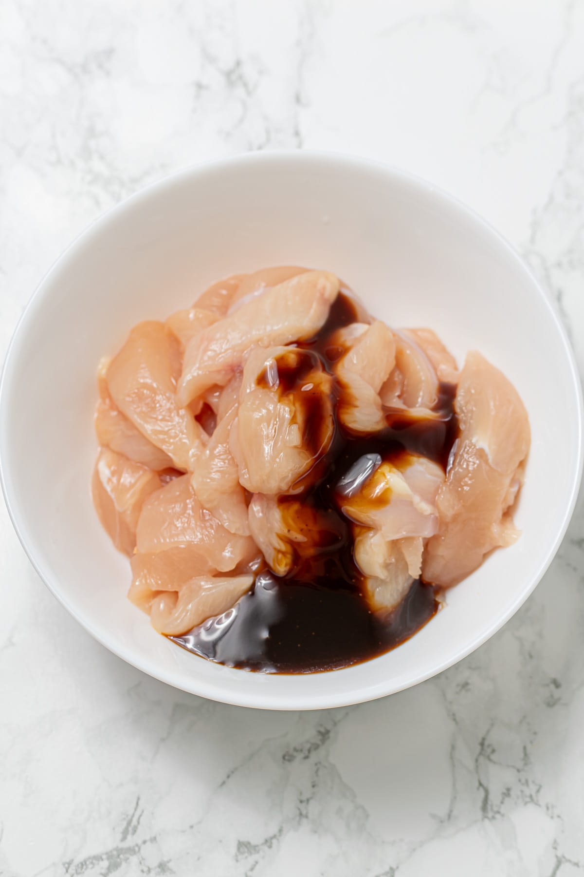 Raw chicken slices in a white bowl with teriyaki sauce poured on top.