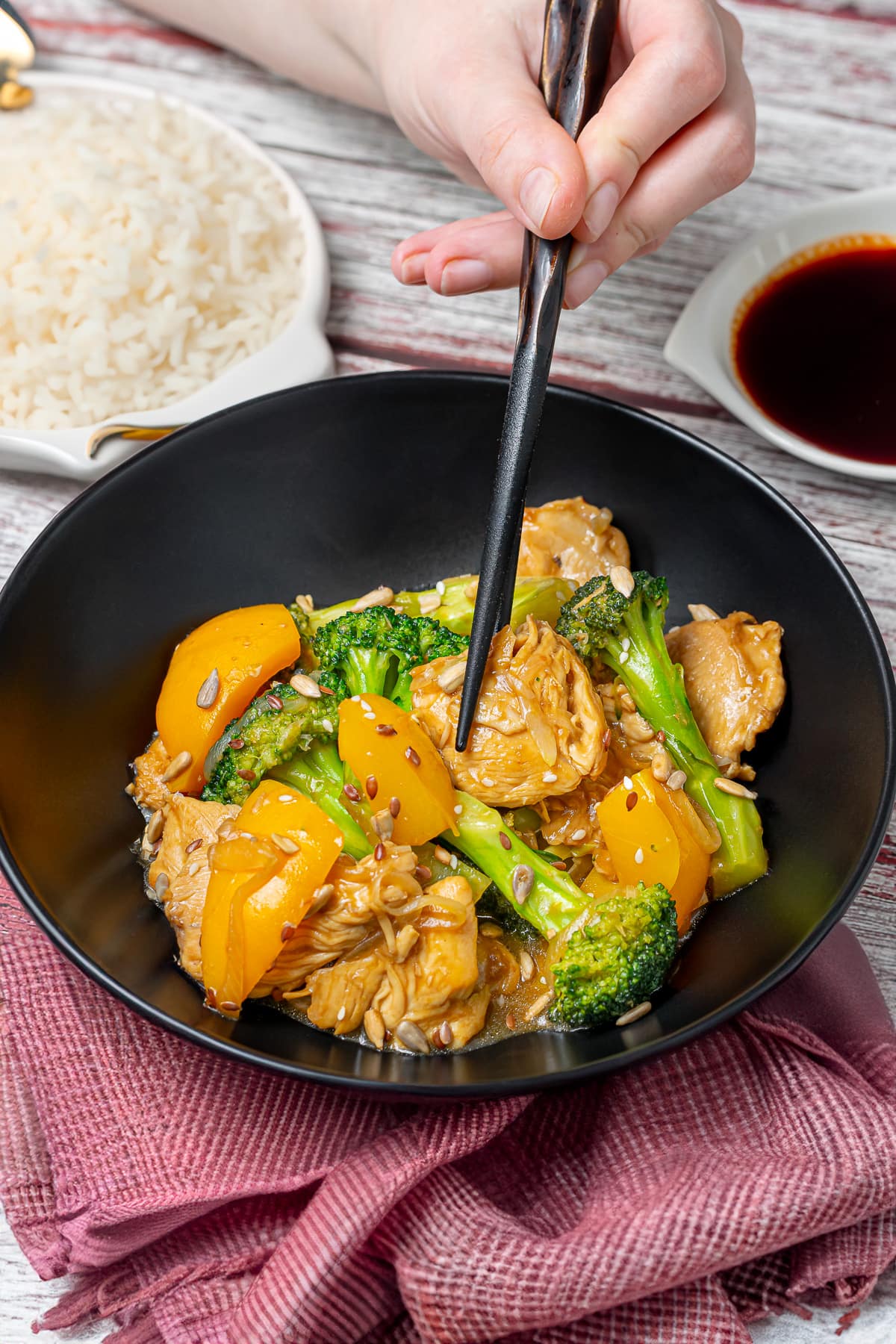 A bowl of teriyaki chicken stir-fry with broccoli and bell peppers, being picked up with chopsticks, over a rustic wooden table with cooked rice and soy sauce in the background.