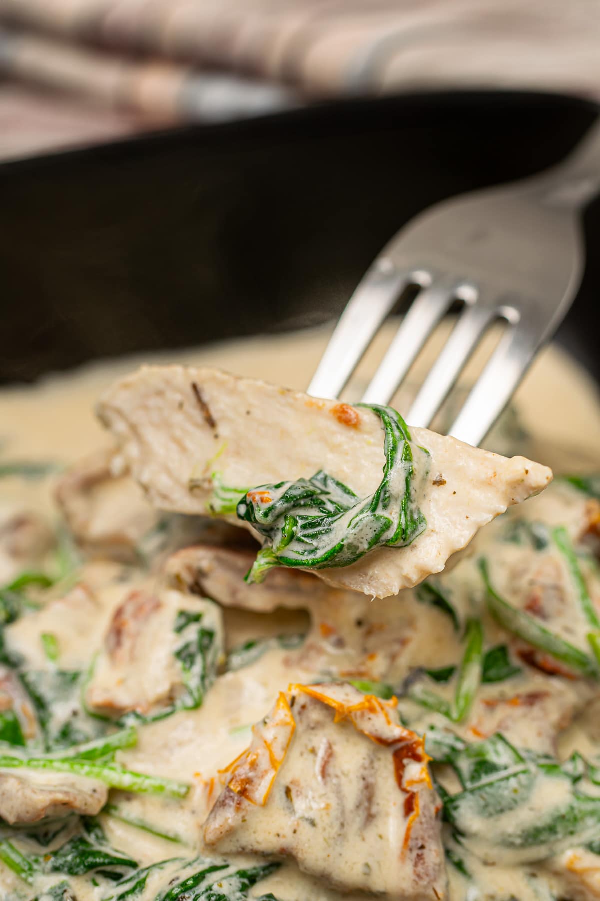 A close-up of a single piece of Tuscan chicken on a fork, showing the creamy sauce and wilted spinach on the chicken.