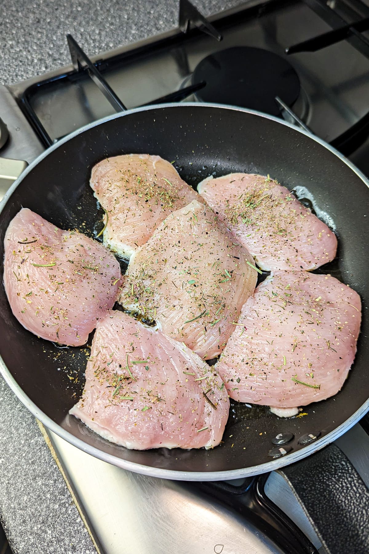Raw chicken breasts seasoned with herbs frying in a skillet on a stove.